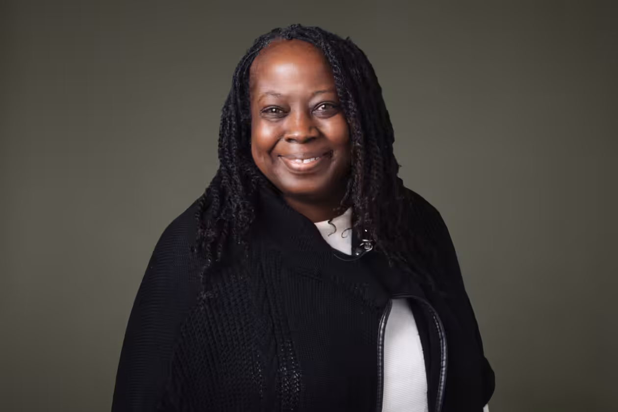 A smiling woman with long, braided hair wears a black sweater over a white top. She stands in front of a plain olive green background.