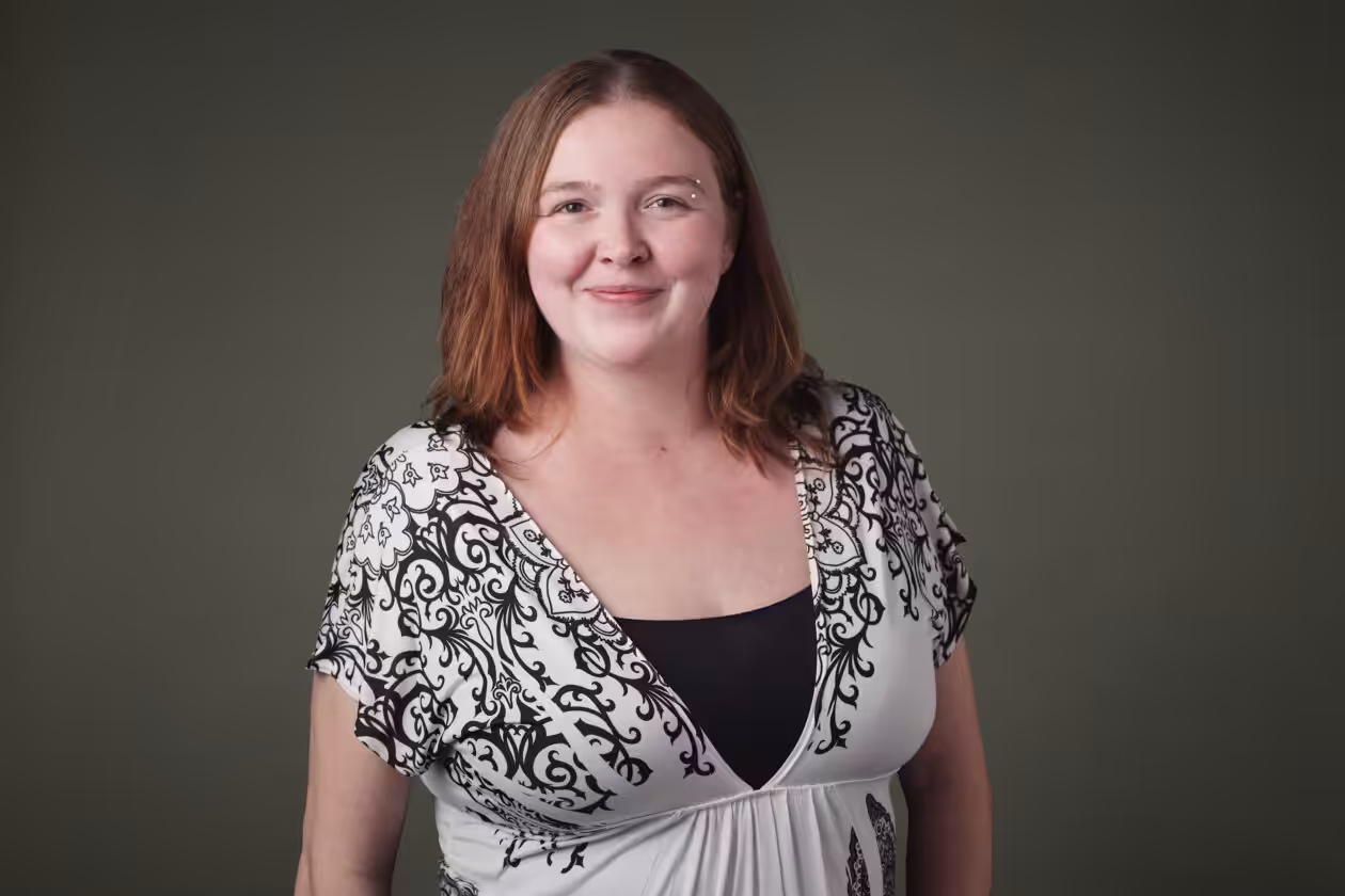 A woman with light brown hair smiles at the camera. She is wearing a white and black patterned blouse over a black top, standing in front of a plain dark green background.