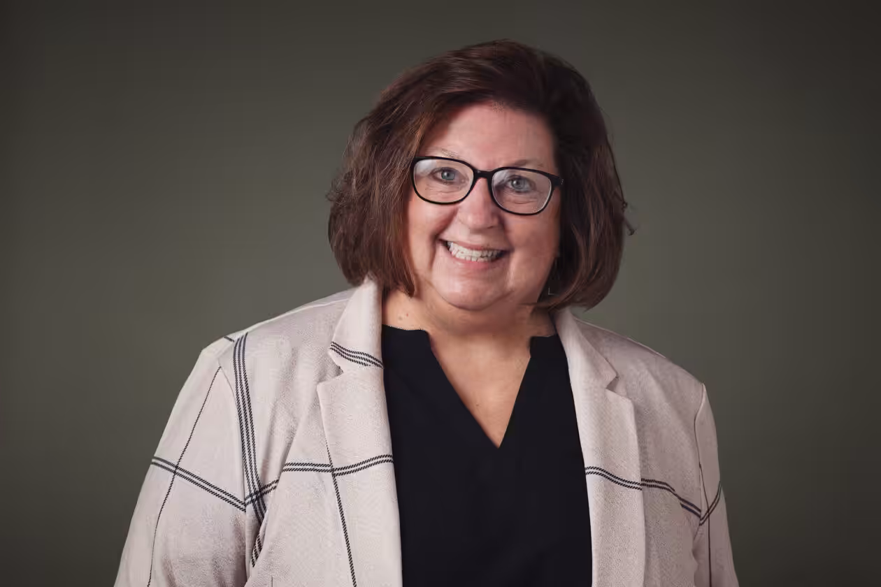 A woman with shoulder-length brown hair, glasses, and a beige plaid blazer over a black top smiles at the camera against a plain dark green background.