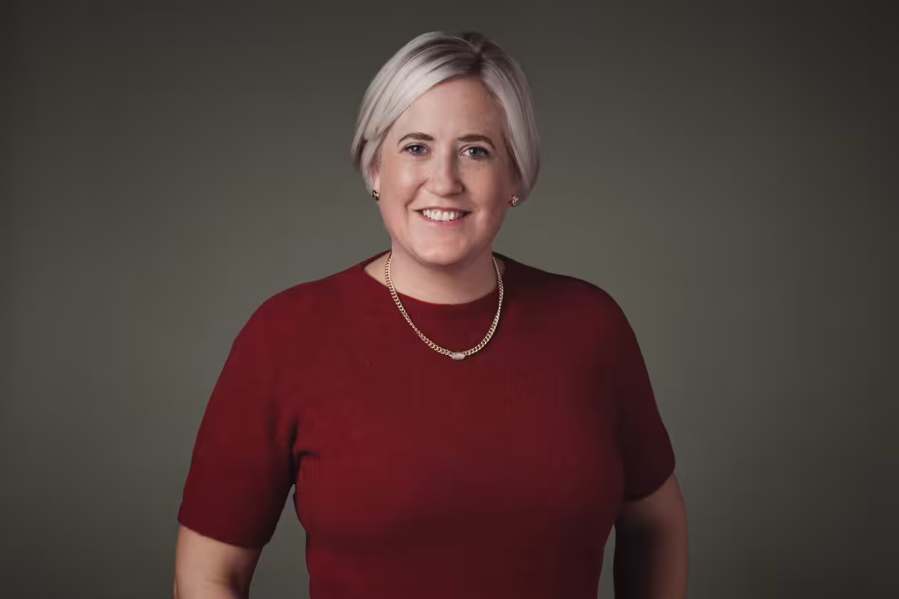 Smiling person with short blond hair wearing a red top and a gold necklace, posing in front of a plain olive-green background.