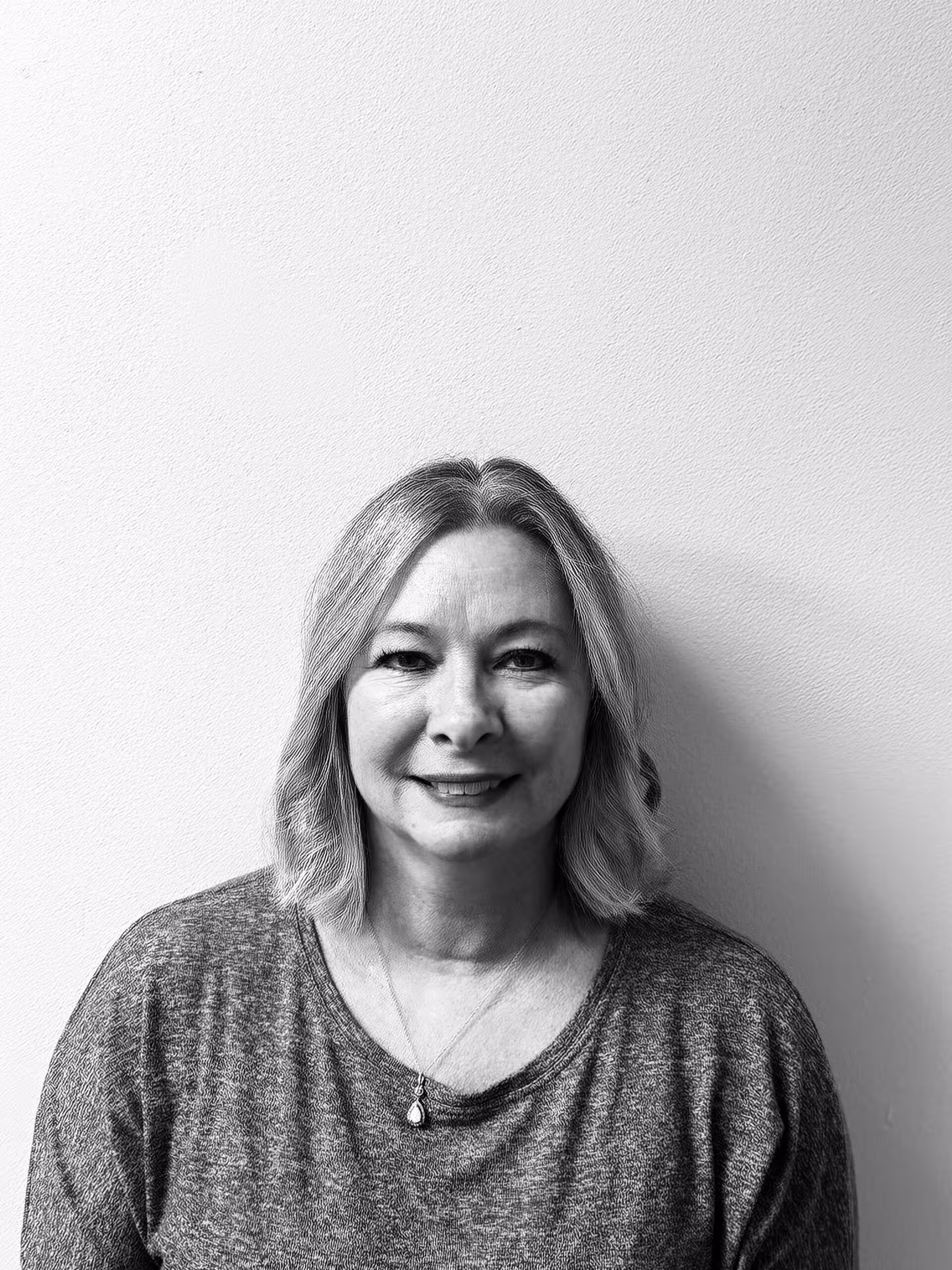 Black and white photo of a woman with shoulder-length light hair, wearing a gray top and a necklace, smiling slightly while sitting against a plain light-colored wall.
