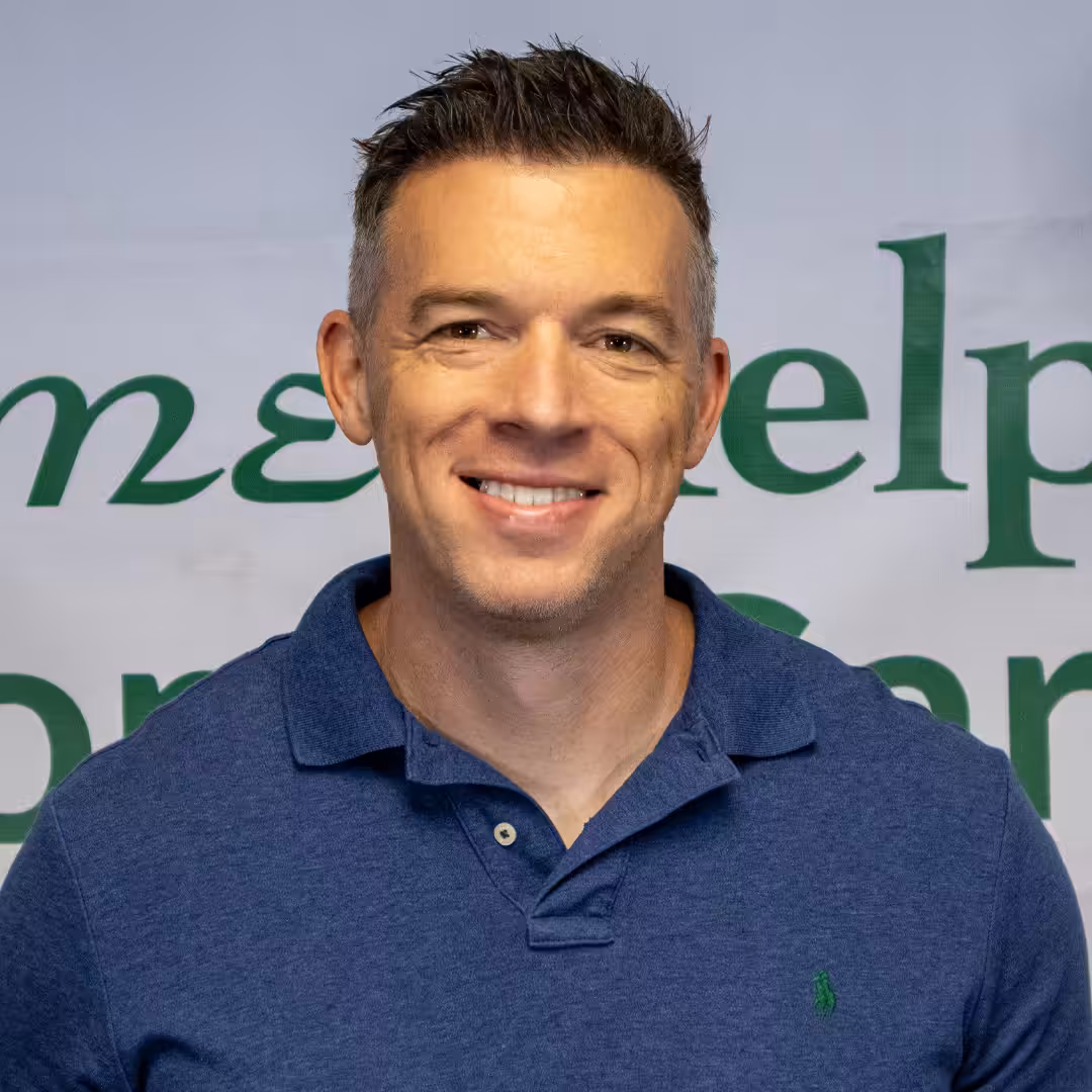 A man with short dark hair wearing a blue polo shirt smiles at the camera, standing in front of a sign with partially visible green text.