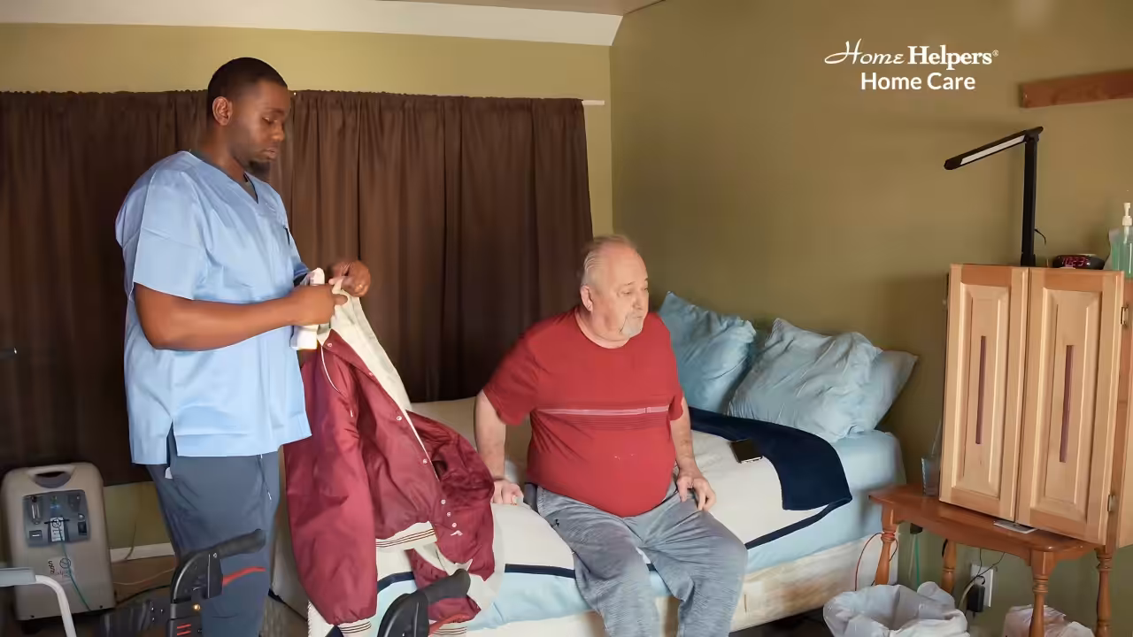 A caregiver in blue scrubs assists an elderly man sitting on a bed in a bedroom, preparing to help him put on a maroon jacket. The room is softly lit, with medical equipment and a wooden cabinet visible.