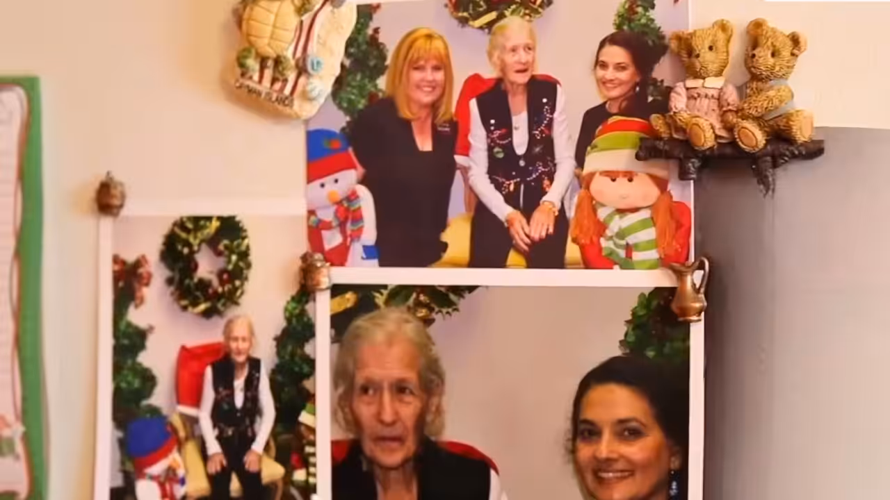 Photos of three people, including an elderly woman, are displayed on a wall decorated with Christmas-themed ornaments, a teddy bear, and festive plush dolls. Christmas wreaths and holiday decor appear in the background.