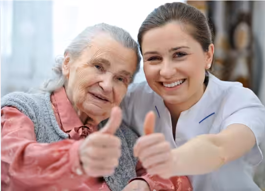 An elderly woman and a younger woman smile at the camera, both giving thumbs up. The elderly woman wears a grey vest, and the younger woman is dressed in a white uniform, suggesting a caregiving or healthcare setting.