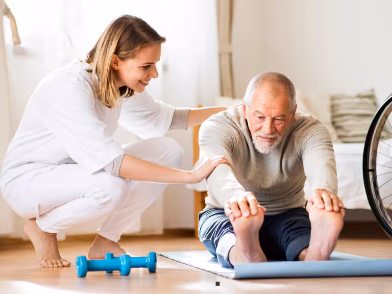 A smiling woman assists an older man as he stretches forward while sitting on a yoga mat in a bright room; a pair of blue dumbbells and a wheelchair are nearby.