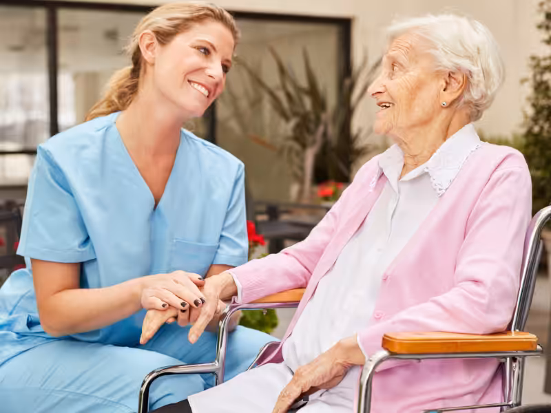 A nurse in blue scrubs smiles and holds hands with an elderly woman in a wheelchair, who is wearing a pink cardigan. They are sitting together outside in a garden area, sharing a warm moment.
