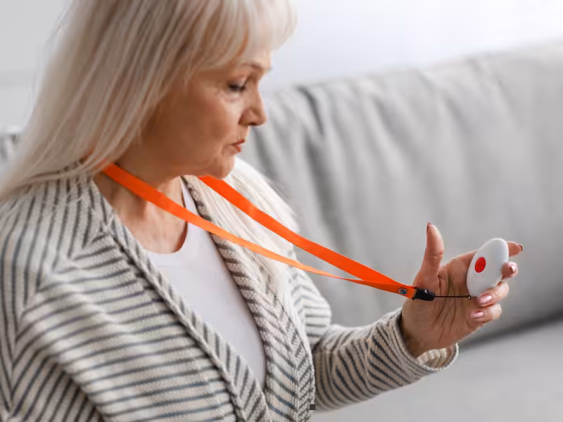 An older woman sits on a couch, holding a white medical alert device with a red button, attached to an orange lanyard around her neck.