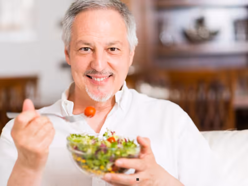 A smiling older man in a white shirt sits indoors, holding a fork with a cherry tomato and a glass bowl of fresh salad.
