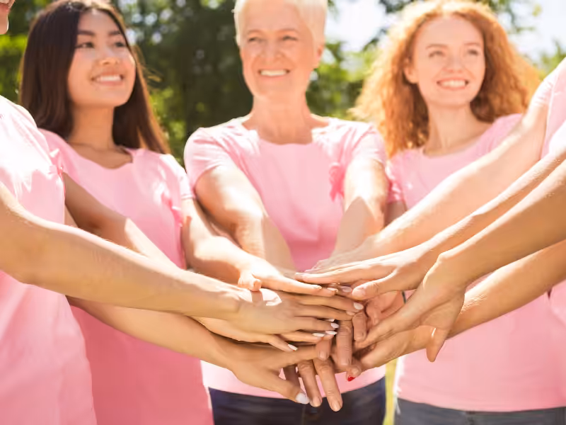 A group of diverse women wearing pink shirts stand outdoors, smiling and stacking their hands together in solidarity, with greenery in the background.