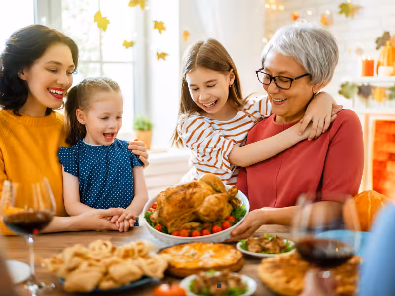 Three generations of women and girls smile and gather around a table, admiring a roast turkey dish at a festive family meal, with autumn decorations in the background.