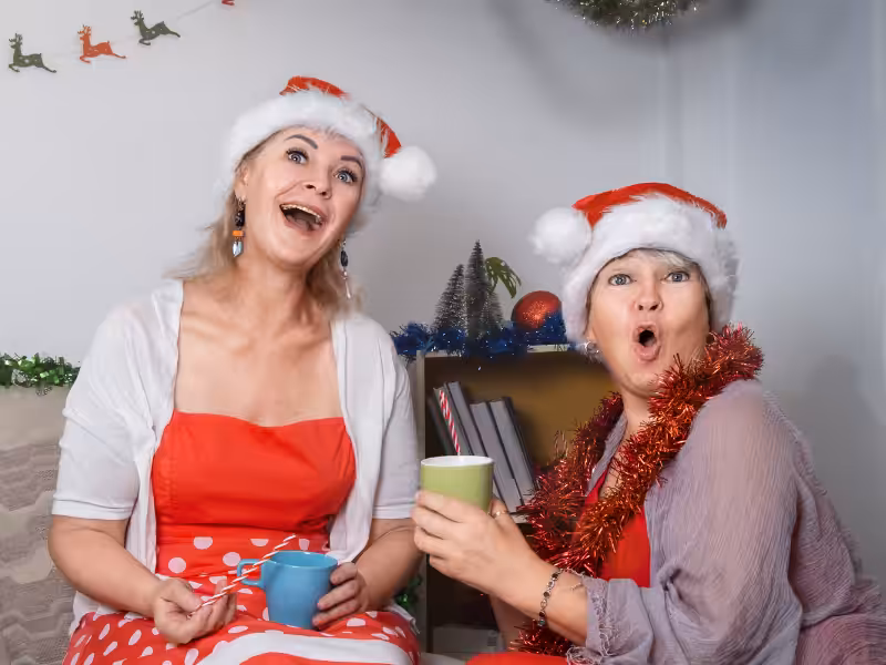 Two women wearing Santa hats and festive red outfits sit on a couch, smiling and looking surprised, holding colorful mugs. Holiday decorations and tinsel are in the background.