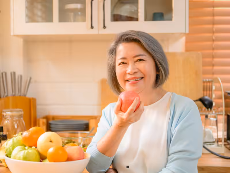 A smiling older woman with gray hair, wearing a light blue cardigan, sits in a kitchen holding a red apple. A bowl of assorted fruits is on the table in front of her. The kitchen background includes shelves and utensils.