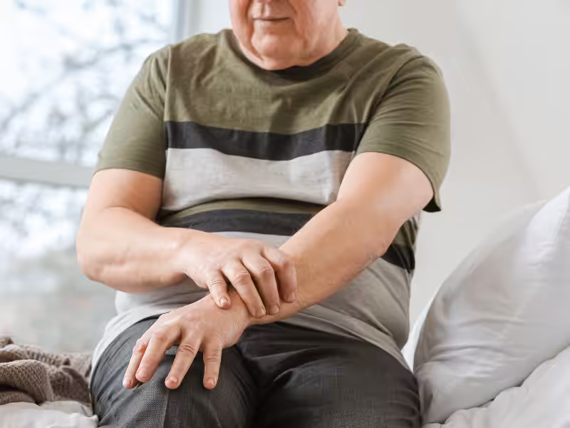 An older adult sitting on a bed, holding and rubbing their wrist with a pained expression, possibly experiencing discomfort or joint pain.