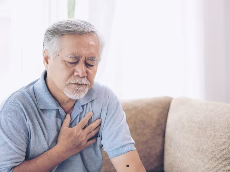 An older man with gray hair and a beard sits on a couch, wearing a light blue shirt. He looks uncomfortable, with his eyes closed and one hand placed on his chest, suggesting chest pain or discomfort.