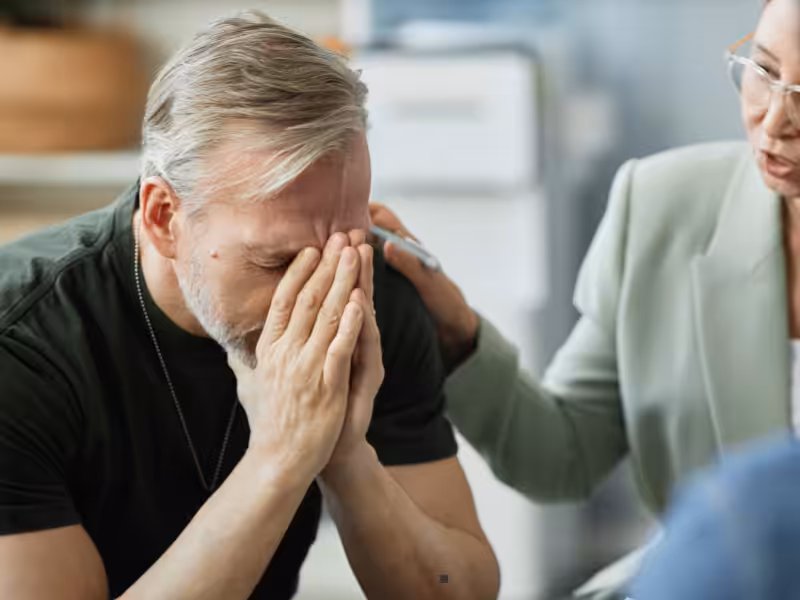 A man sits with his hands covering his face, appearing distressed, while another person beside him gently touches his shoulder in a comforting gesture.