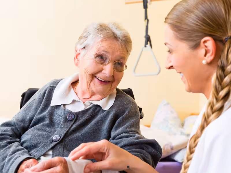 An elderly woman in a wheelchair smiles warmly at a young female caregiver, who is gently holding her hand in a bright, comfortable room.