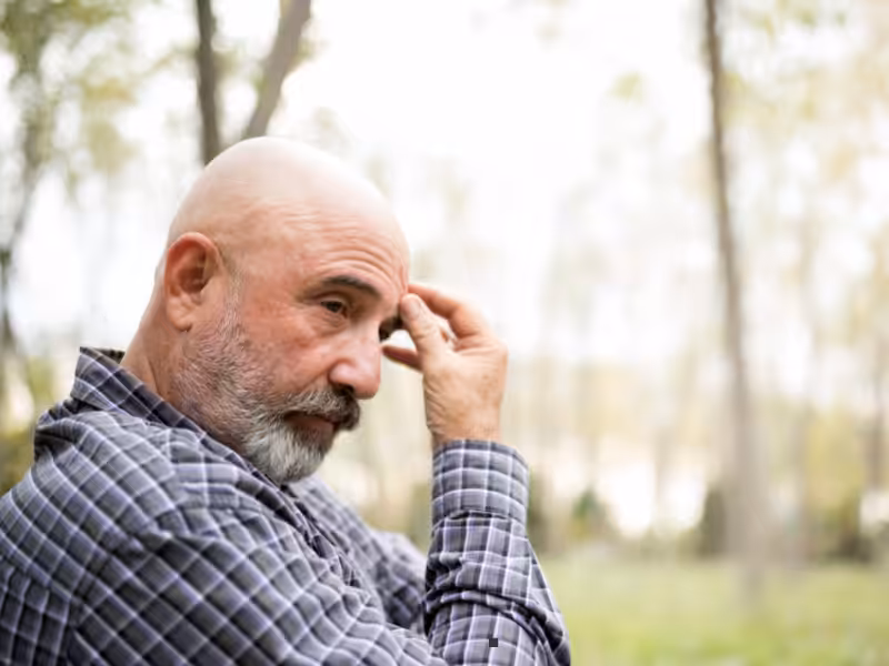 A bald man with a gray beard, wearing a plaid shirt, stands outdoors in a forested area, touching his forehead while looking thoughtful. The background is blurred with trees and soft natural light.