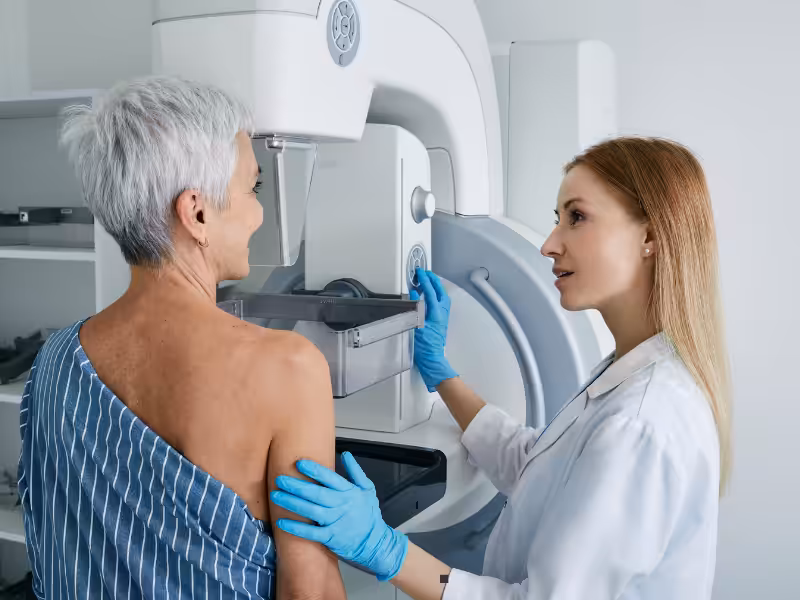 A healthcare professional assists a woman with short gray hair during a mammogram procedure, guiding her at a medical imaging machine in a clinical setting.