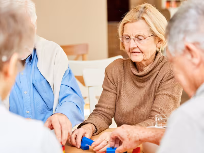 Four older adults are seated around a table, concentrating on a colorful block game together. They are engaged and interacting, suggesting a social and recreational activity in a bright indoor setting.