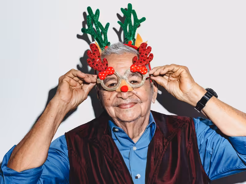Smiling older man wearing a blue shirt, maroon vest, and festive reindeer antler glasses with a red nose, posing playfully against a plain white background.