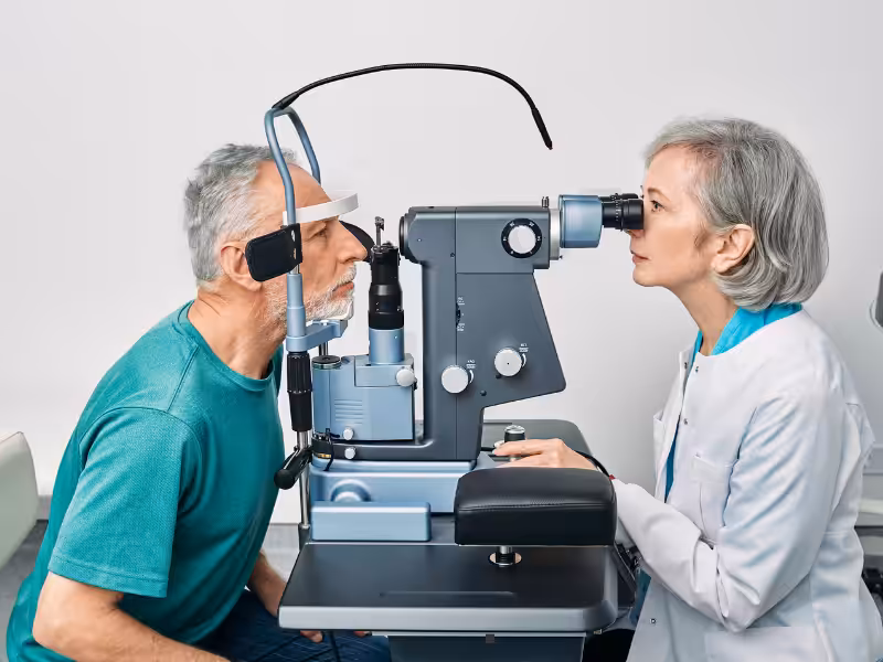 An older man is having his eyes examined by a female optometrist using a slit lamp in a medical office. The man is seated with his chin on the chin rest while the doctor looks through the device.