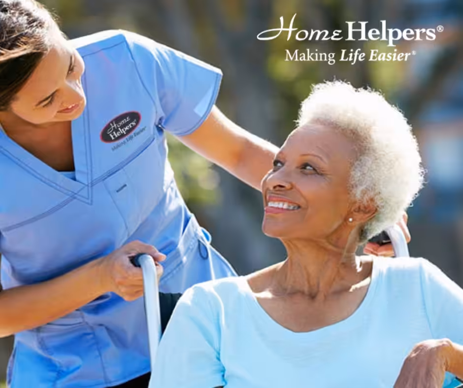 A smiling elderly woman sits in a wheelchair outdoors while a caregiver in blue scrubs stands beside her, smiling warmly. The Home Helpers logo and the phrase Making Life Easier appear in the top right corner.