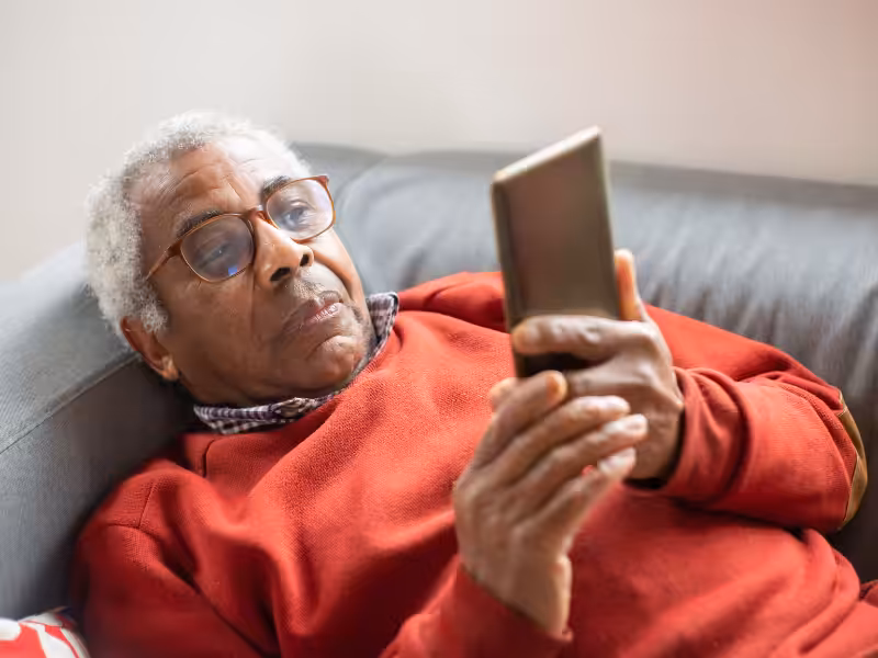 An elderly man with gray hair and glasses, wearing a red sweater, is lying on a couch and holding a smartphone, looking at the screen intently.