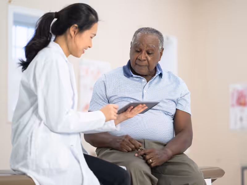 A female doctor shows a digital tablet to an older male patient in a medical exam room, discussing information together.
