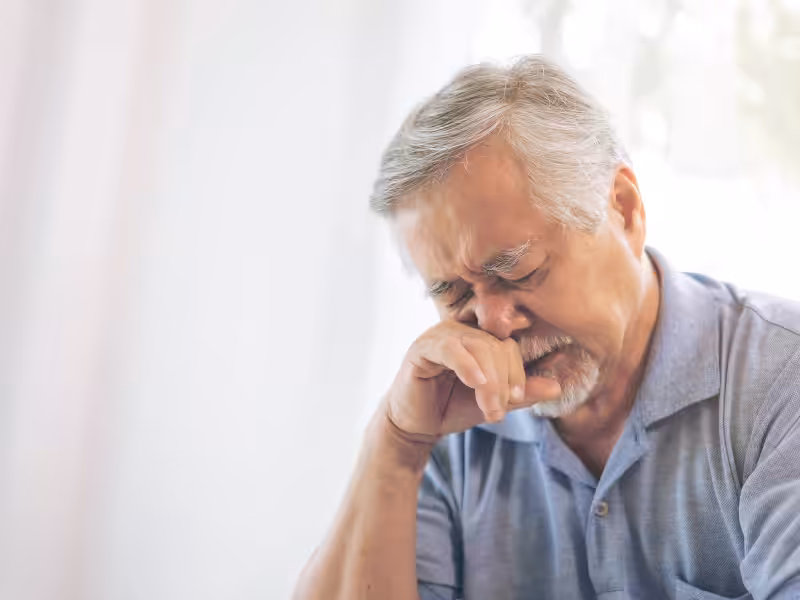 An elderly man with gray hair and a beard, wearing a blue polo shirt, sits with his eyes closed and his hand covering his mouth, appearing sad or deep in thought.