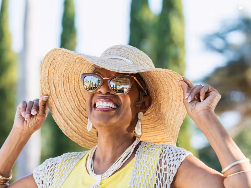 Smiling woman wearing sunglasses and a large straw hat, holding the brim with both hands, standing outdoors with trees and blue sky in the background.