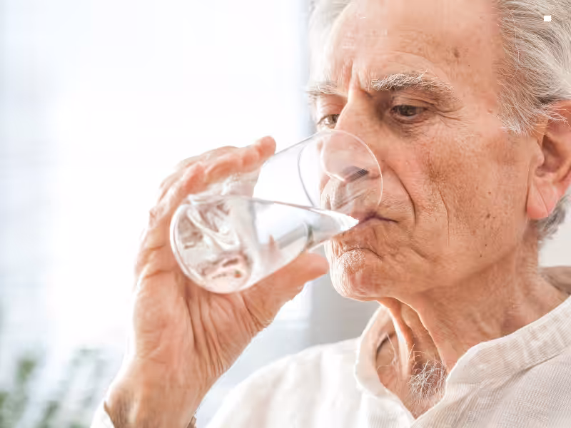 An elderly man with gray hair drinks a glass of water, wearing a light-colored shirt, in a bright, softly lit setting.