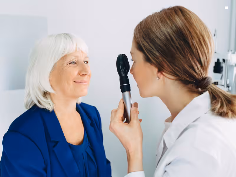 An eye doctor examines an older woman’s eyes using an ophthalmoscope during an eye checkup. The woman has white hair and is smiling, while the doctor wears a white coat.