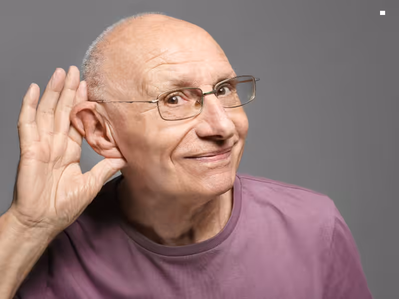 Older man with glasses wearing a purple shirt, smiling and holding his hand to his ear in a listening gesture against a gray background.