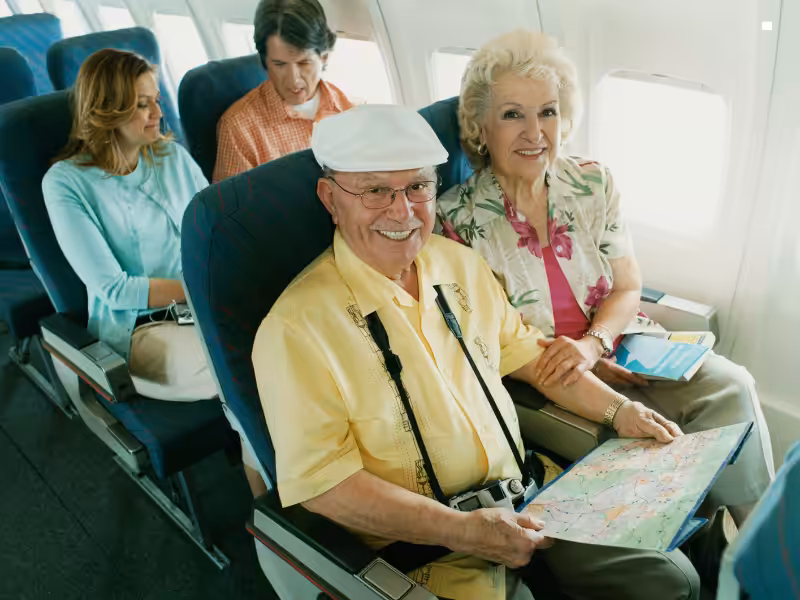 An elderly couple smiles while sitting on an airplane, the man holding a map and wearing a camera around his neck. Two other passengers sit behind them, looking at travel documents.