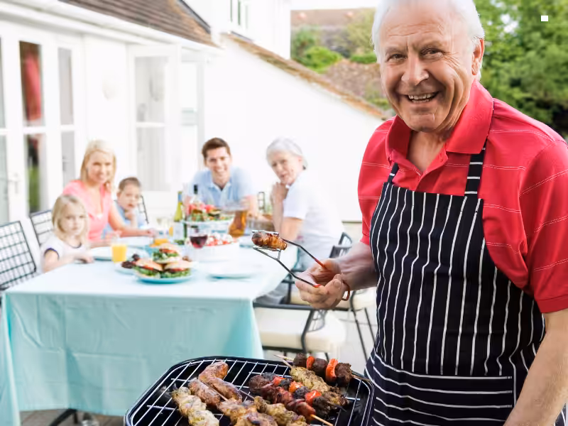 An older man in a striped apron grills skewers outdoors, smiling at the camera, while a family of four sits at a table behind him enjoying a meal in a sunny backyard.
