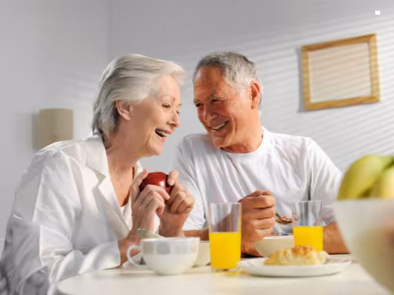 An elderly couple sits at a breakfast table, smiling and laughing. The woman holds an apple, while the man eats with a spoon. There are two glasses of orange juice and a plate of food on the table.