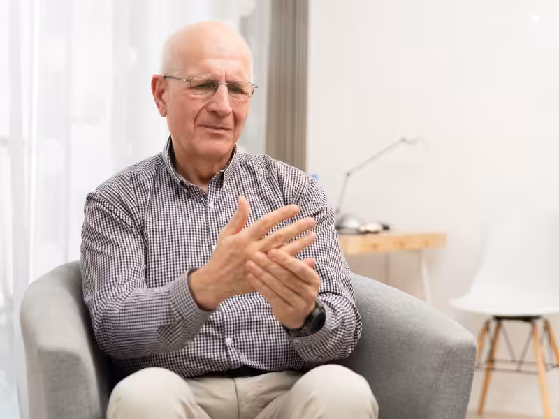 An older man with glasses sits in an armchair, holding his right hand with his left as if experiencing discomfort. He wears a checked shirt and looks thoughtful. A desk and chair are in the background of the bright room.