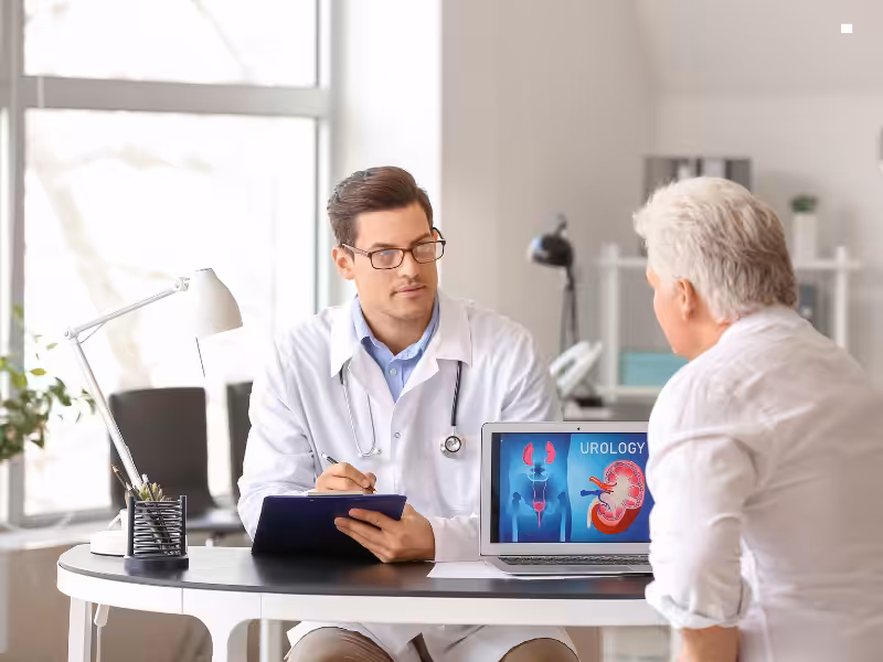 A doctor in a white coat consults with an older male patient in an office. A laptop on the desk displays urology-related images of kidneys and the urinary system.