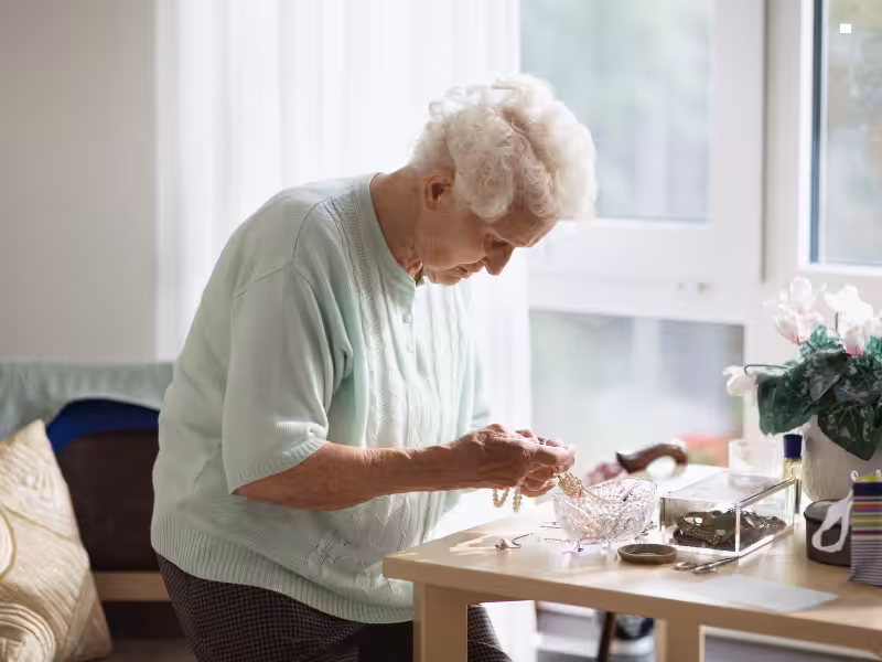 An elderly woman with short, curly white hair and a light green sweater stands by a table, looking down as she organizes jewelry in a well-lit room with large windows and a potted plant nearby.