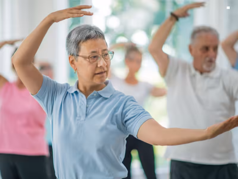 A group of seniors participate in a fitness or tai chi class indoors, focusing on slow, graceful movements. The woman in the foreground wears glasses and a light blue shirt, leading the motion with her arms raised.