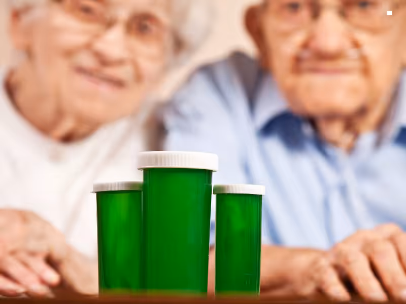 Two elderly people sit together in the background, with three green prescription pill bottles with white caps placed prominently in the foreground. The focus is on the pill bottles.