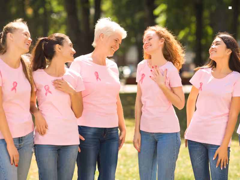 Five women wearing pink T-shirts with pink ribbons stand together outdoors, smiling and talking, supporting breast cancer awareness. Trees and greenery are visible in the background.