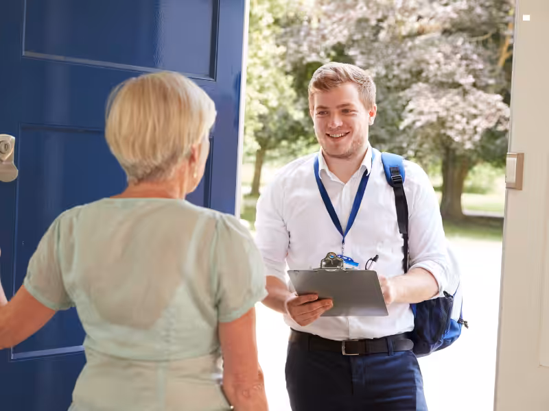 A smiling young man with a clipboard and lanyard stands at an open door, speaking to an older woman with short gray hair, who is facing him. Trees and greenery are visible outside.