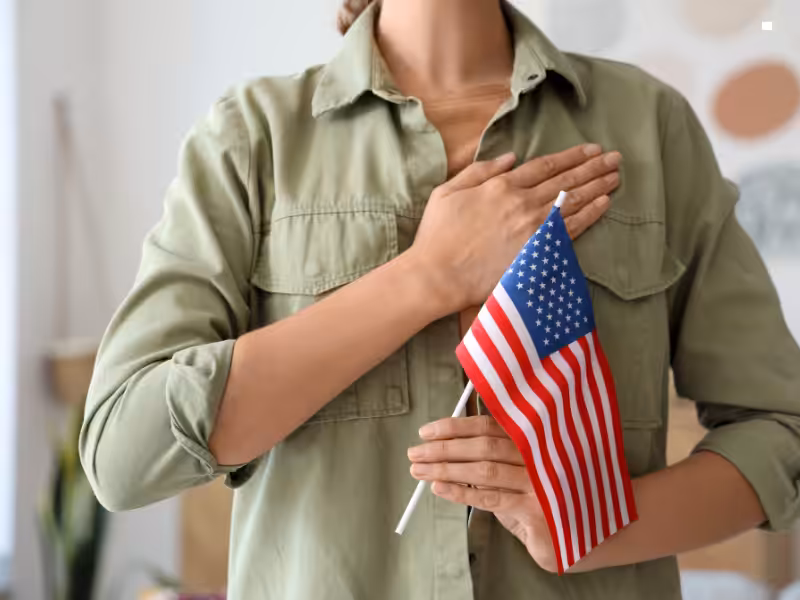 A person wearing a green shirt holds a small American flag in one hand and places their other hand over their heart, symbolizing patriotism or respect for the United States.