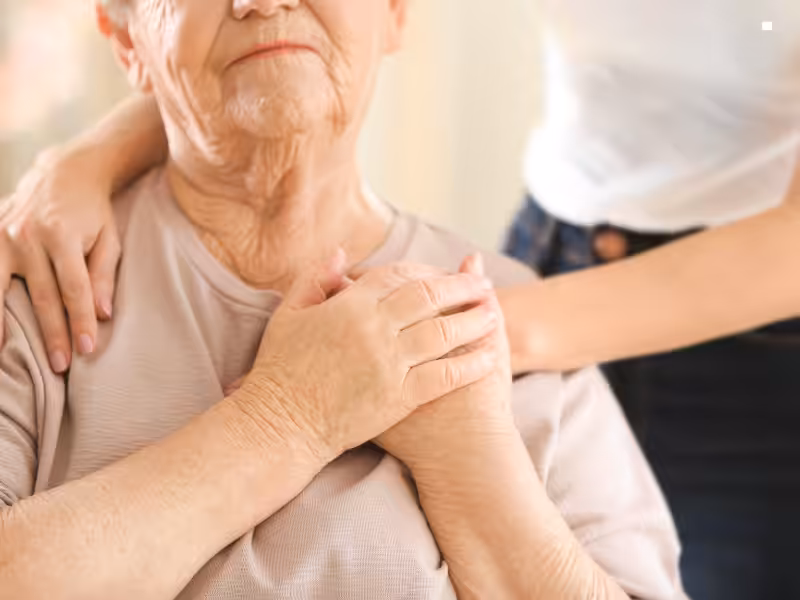 An elderly person sits with hands clasped over their chest as another person stands behind, gently resting their hands on the elder’s shoulders, offering comfort and support.