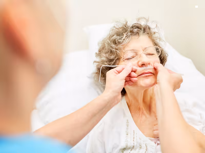 An older woman with gray hair lies in a hospital bed while a healthcare worker gently inserts a nasal cannula for oxygen support. The woman’s eyes are closed, and the setting appears clinical.
