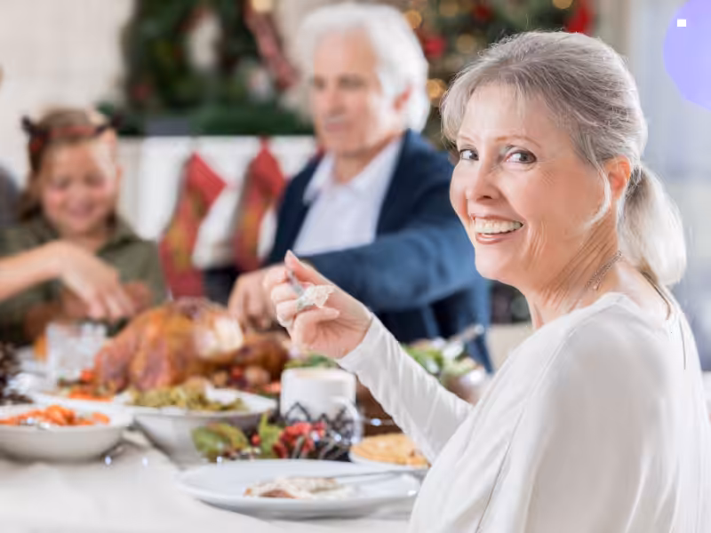 Smiling older woman sitting at a festive dining table, looking at the camera, with family members and holiday food, including a roast turkey, in the background.