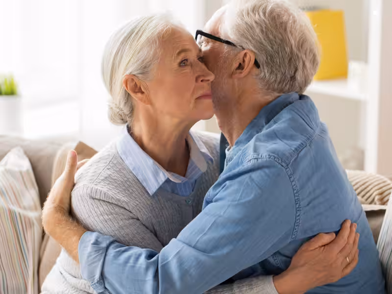 An elderly couple sits on a couch, hugging. The man gently kisses the woman on the cheek, while she looks thoughtful and slightly distant. They are in a bright, cozy room with soft lighting.