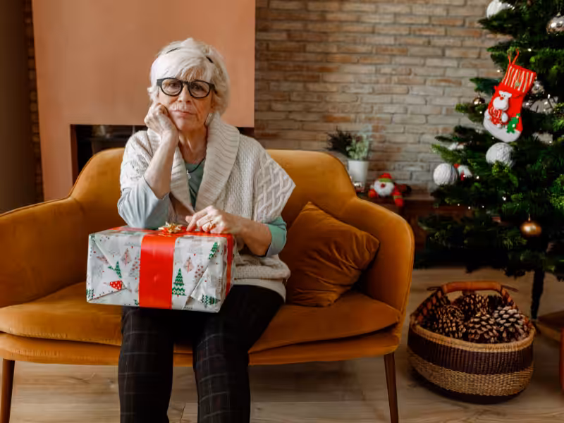 An elderly woman with glasses sits on a couch holding a wrapped Christmas present, looking pensive. A decorated Christmas tree and a basket of pine cones are beside her in a cozy living room.